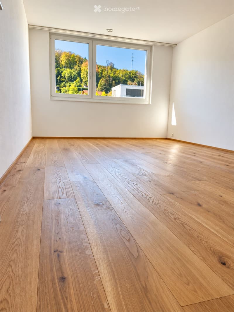Empty room with light wooden flooring and white walls, featuring large windows with a view of greenery outside.