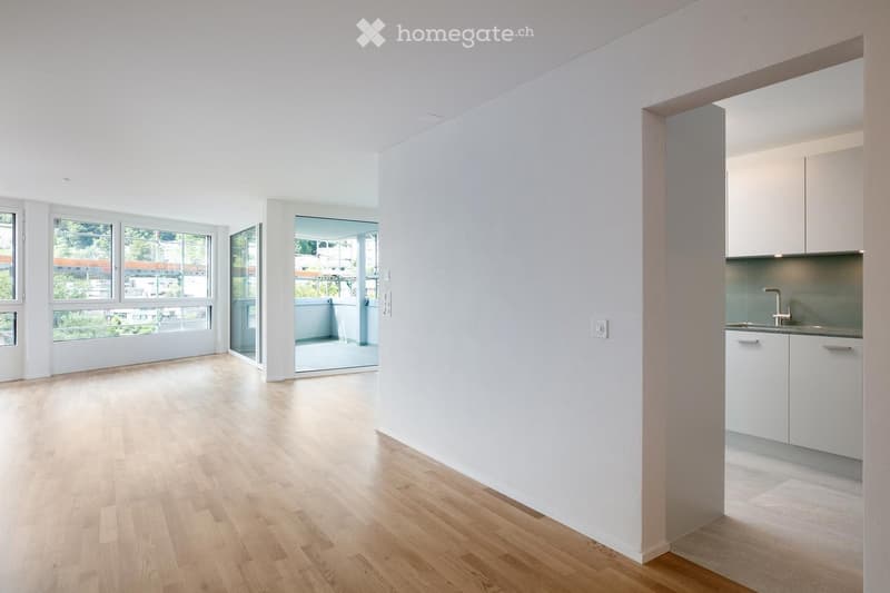 Bright, unfurnished room with light wooden flooring, white walls, large windows, and a small kitchen area with white cabinets and a sink.