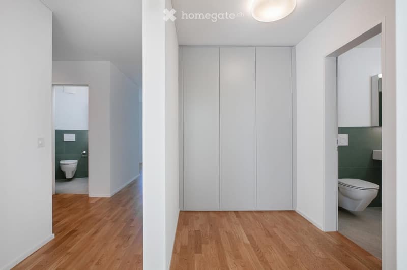A modern, minimalist hallway with light wooden flooring, white walls, and built-in white closet units. Two bathrooms are visible at the ends of the hallway, each with a toilet and green tiled walls.