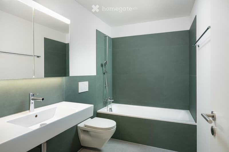 Modern bathroom with a white sink, chrome faucet, toilet, bathtub, and showerhead. Green tiles on the walls, white cabinetry, and a large mirror above the sink.