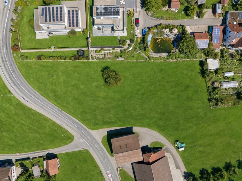 Aerial view of a residential area with multiple houses, solar panels, a pond, and surrounding greenery.