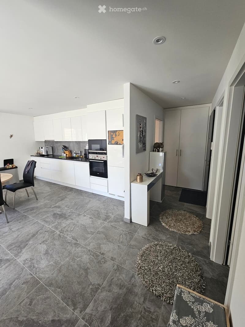Modern kitchen with white cabinets, stainless steel appliances, and a dining area with a round table and chairs. The floor is tiled in grey marble, and there are two small round rugs in the seating area.
