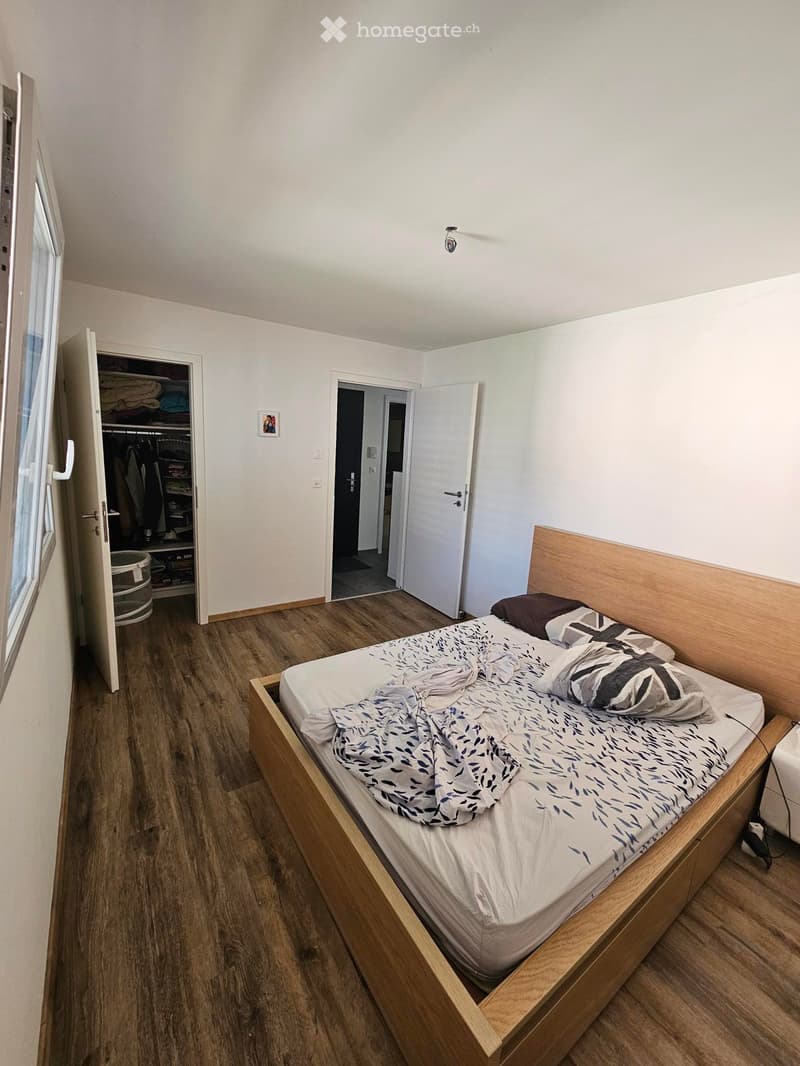 A tidy bedroom with a wooden bed, patterned bedding, a bedside table, an open wardrobe, and a picture frame on the wall.