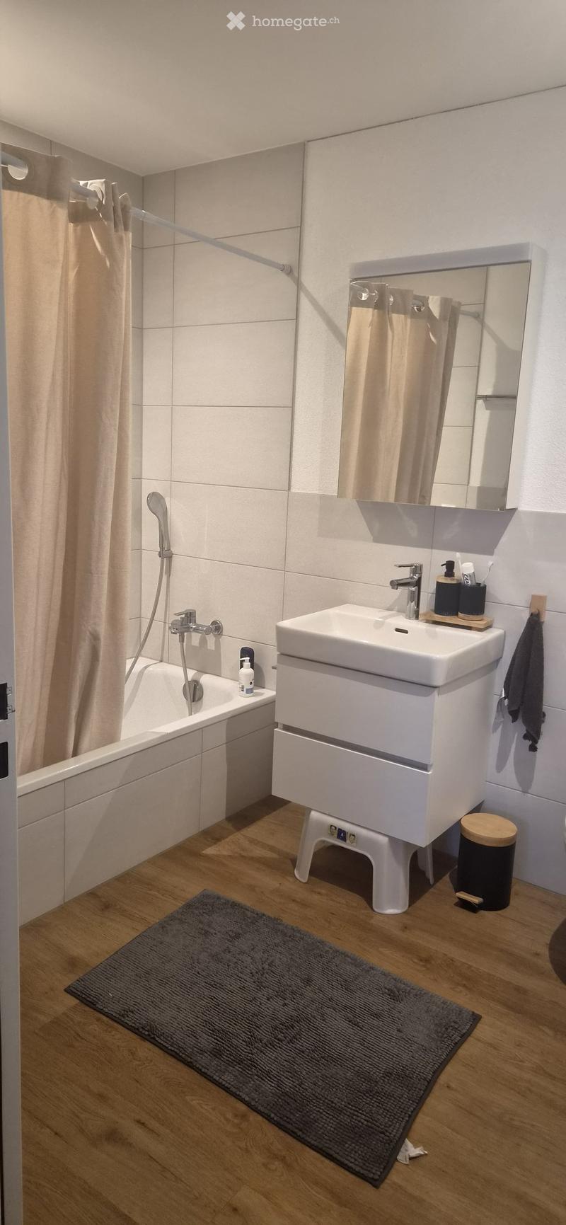 Modern bathroom with a white sink, bathtub, mirror, and wooden floor. Includes a grey bath mat, a small stool, and a toilet.