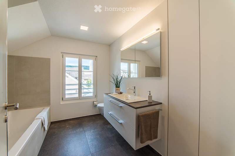 Modern bathroom with a bathtub, sink, and toilet. Features a large mirror with built-in lighting, a window with a view, and a small potted plant on the sink counter.