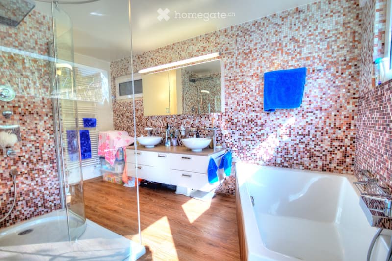 Modern bathroom with a bathtub, double sink, large mirror, and a glass-enclosed shower. The walls are tiled with a mosaic pattern, and the floor is wooden.