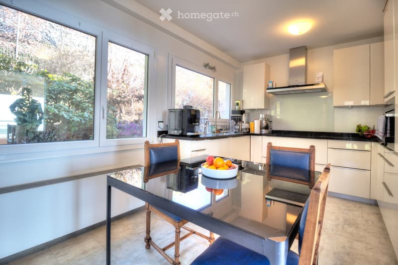 Modern kitchen with white cabinets, a glass dining table with four chairs, a bowl of fruit on the table, and various appliances including a coffee maker and a range hood.