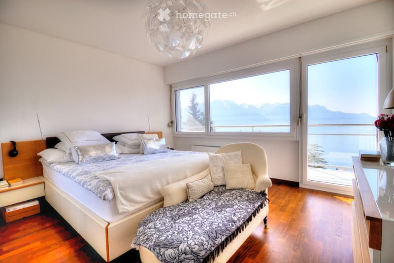 Elegant bedroom with a large bed, patterned throw, decorative pillows, a white armchair, a glass table with a vase of flowers, and a scenic mountain view through large windows.