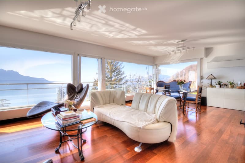 Modern living room with large windows offering a scenic mountain view, featuring a white leather sofa, glass coffee table, and a blue armchair.
