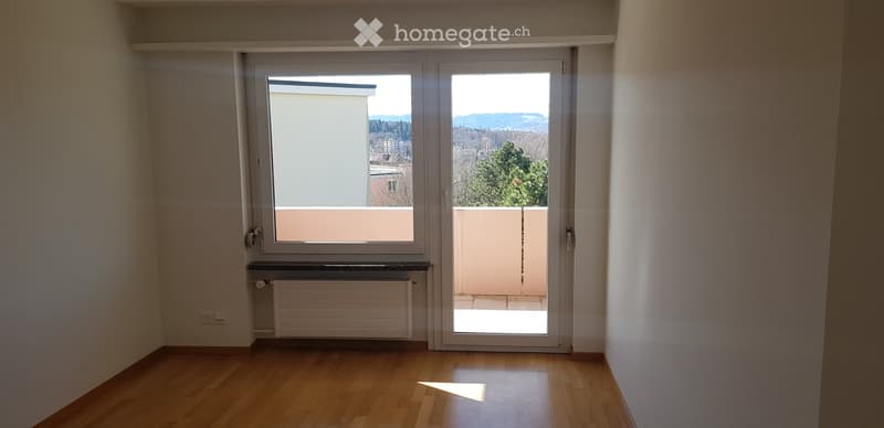 Empty room with wooden flooring, large window, and sliding glass door leading to a balcony. The room has white walls and a radiator below the window.