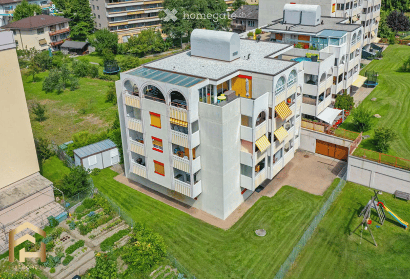 Aerial view of a modern multi-story residential building with balconies, surrounded by green lawns, trees, and playground equipment.