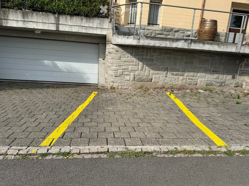 A paved parking area with yellow lines, adjacent to a stone building with a garage and a raised balcony.