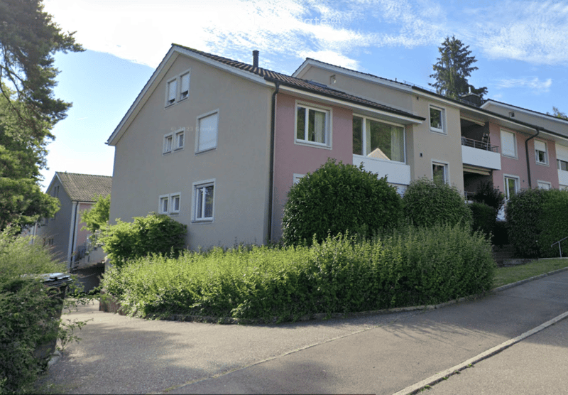Two-story house with beige and pink exterior, multiple windows, and a small balcony on the upper floor. The house is surrounded by greenery and has a driveway in front.