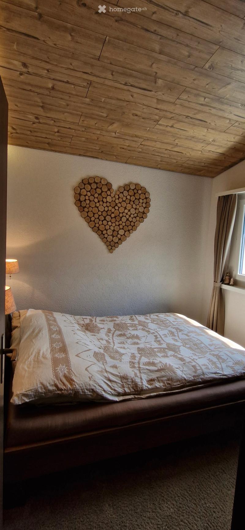 Cozy bedroom with a wooden slat ceiling, a heart-shaped wooden wall decoration, a bed with a patterned duvet, and a nightstand with a lamp.