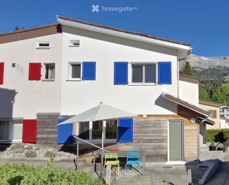 Two-story house with white walls, blue shutters, and a wooden deck area with outdoor furniture and an umbrella.