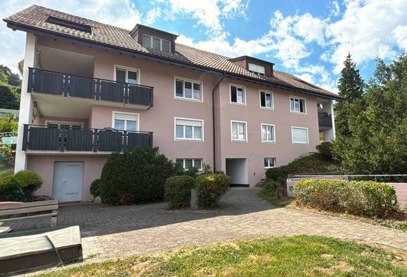 Two-story pink building with multiple balconies, white-framed windows, and a tiled roof. The entrance features a small porch with a door, and the surrounding area includes a paved driveway and landscaped greenery.