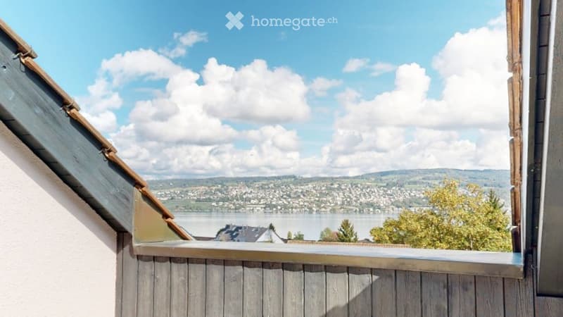Balcony with wooden railing, overlooking a scenic lake and distant town with mountains in the background.
