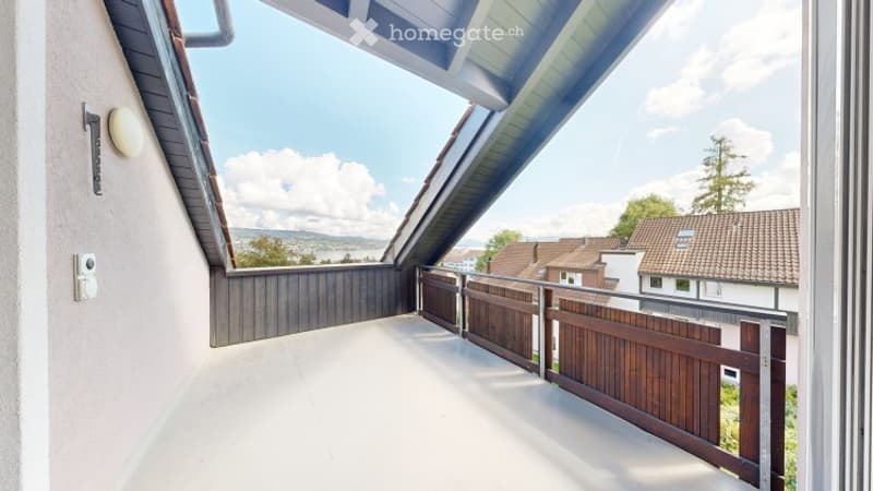 A modern balcony with a wooden railing, white flooring, and a view of the surrounding area with houses and trees.