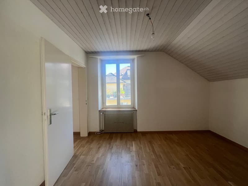 Empty room with wooden flooring, white walls, and a window with a radiator below it. The ceiling is slanted with wooden paneling.