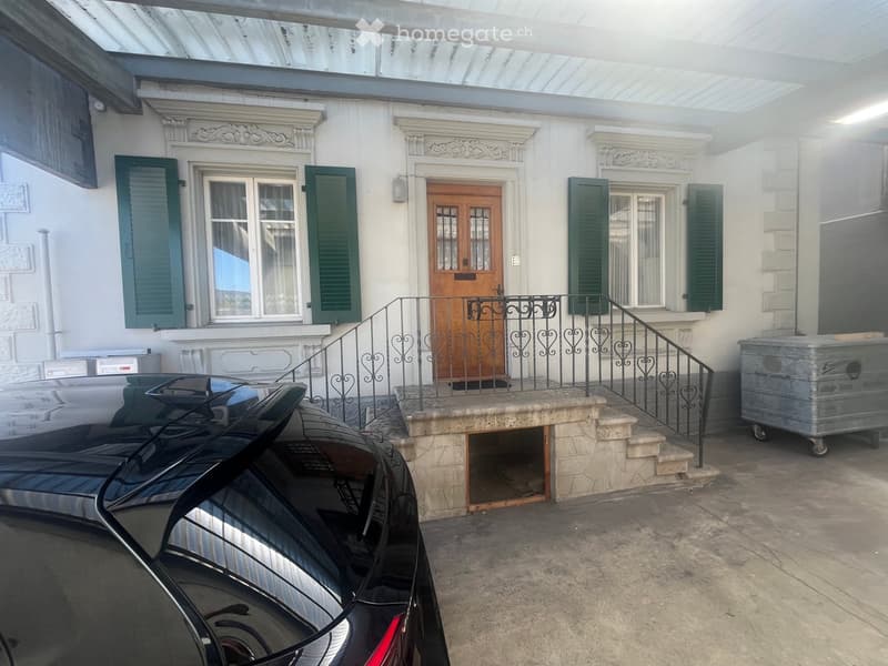 A two-story building with a white facade, green shutters, and a wooden double door. A black car is parked in front, and a metal staircase leads up to the entrance.