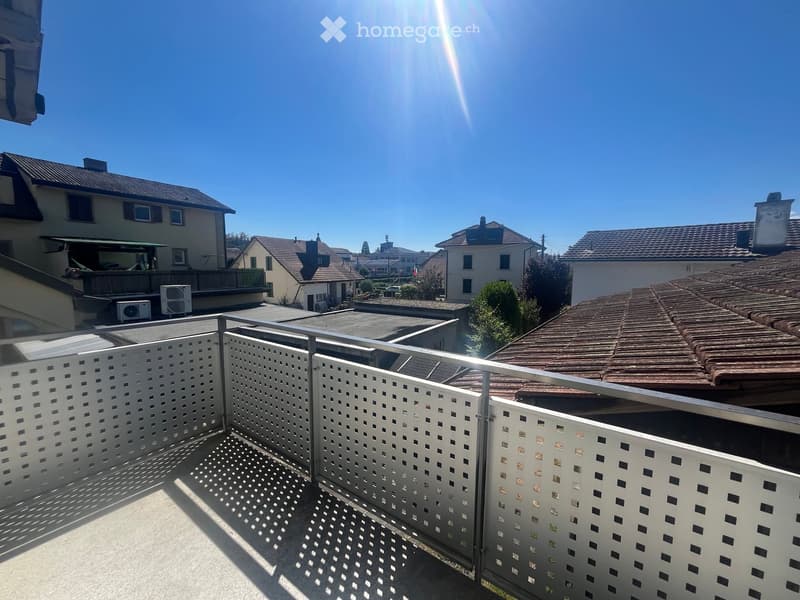 Balcony with white lattice fencing, overlooking a residential area with houses, trees, and a clear blue sky.