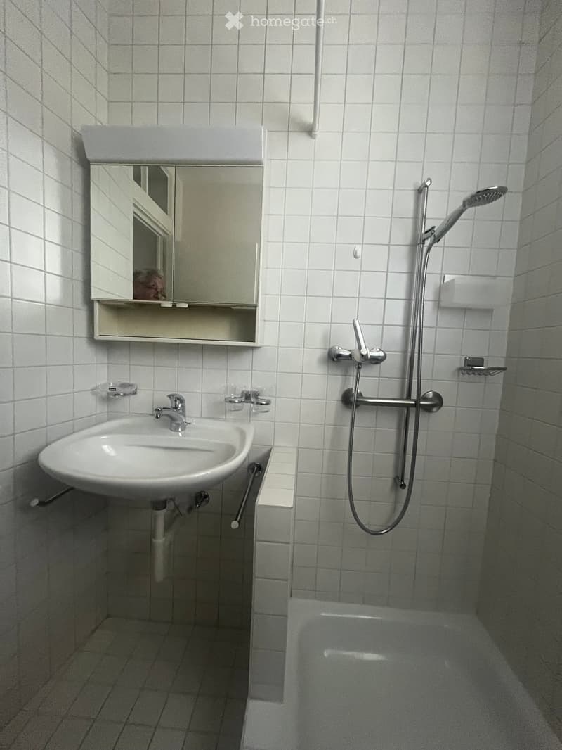 Small bathroom with white tiled walls and floor, featuring a sink with a mirror above it, a bathtub, and a handheld showerhead with a rail.