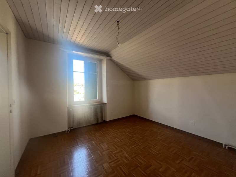Empty room with wooden flooring, white walls, and a single window letting in natural light. The ceiling has a slanted design typical of attic spaces.