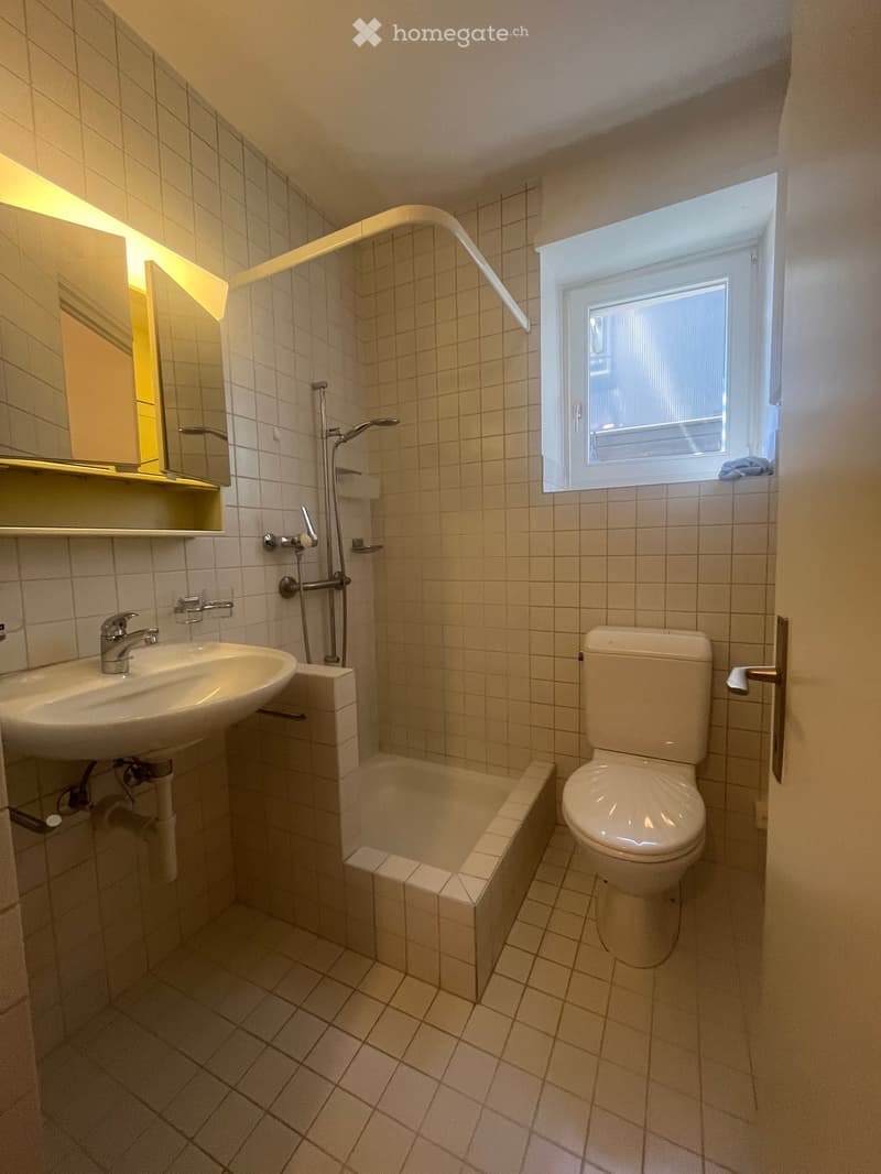 Small bathroom with white tiled walls and floor, featuring a sink, toilet, bathtub, and a window with blinds.