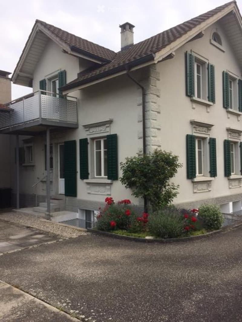 Two-story house with beige walls, green shutters, and a balcony on the upper level. The house features a chimney and is surrounded by a small garden with red flowers and a tree.