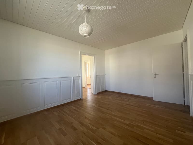 Empty room with light wooden flooring, white walls, and a decorative ceiling light fixture. The room has a doorway leading to another space.