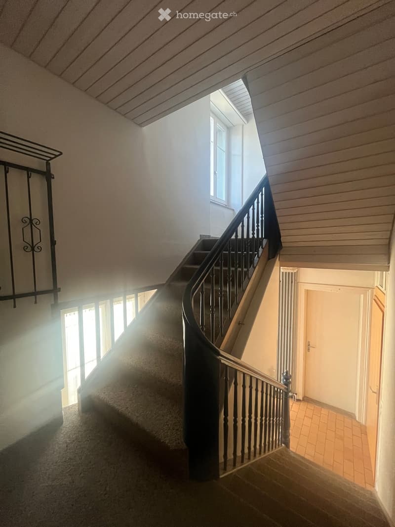 Spiral staircase with wooden steps and dark railing, leading to a room with a tiled floor and white walls. Natural light streams in from a window on the left side.