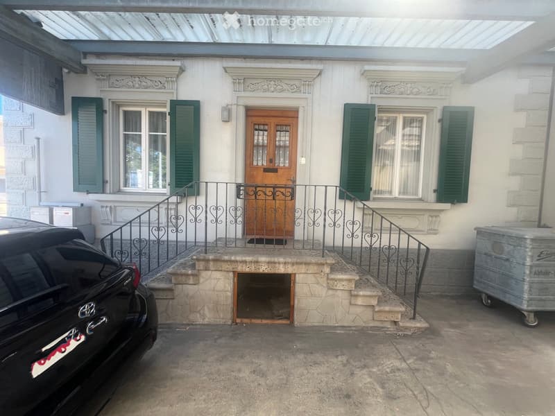 Two-story building with white facade, green shutters, and a wooden double door. A small staircase leads to the entrance, flanked by black wrought-iron railings. A black car is parked on the left, and a large metal container is on the right.