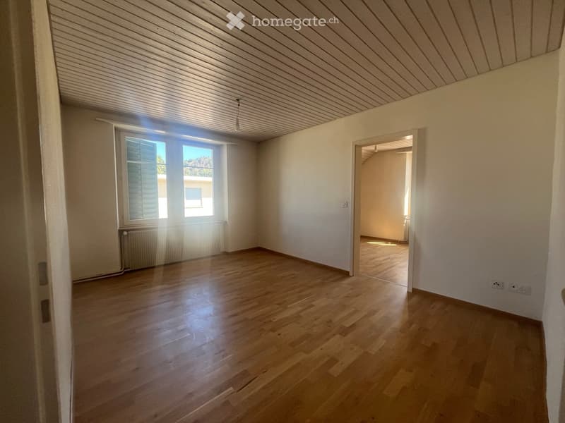 Empty room with wooden flooring, white walls, and a slanted wooden ceiling. Features a window with shutters and a radiator below it, with an open doorway leading to another room.