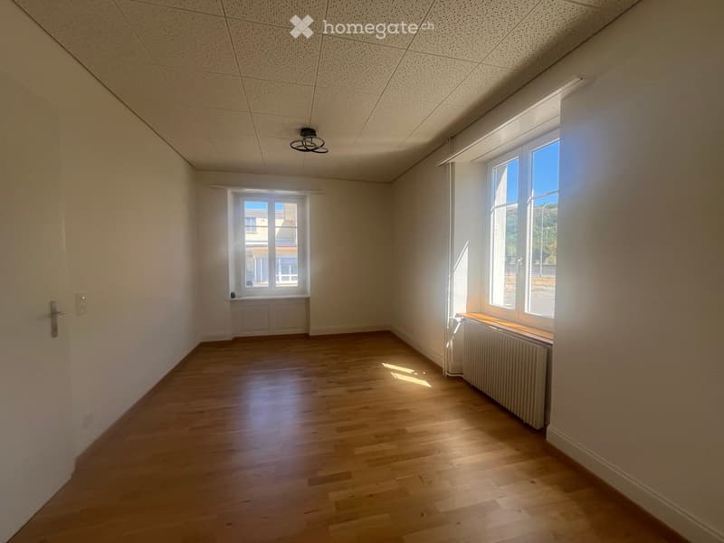 Empty room with wooden flooring, white walls, and two windows allowing natural light. The room features a radiator under one window and a ceiling light fixture.