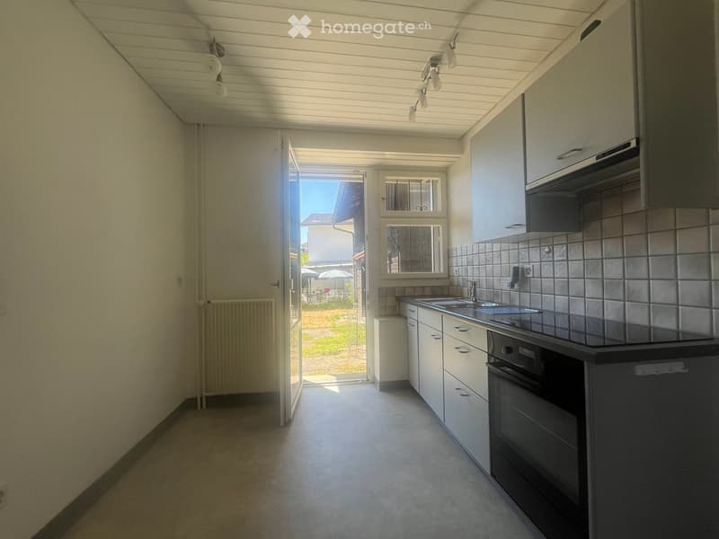 Modern kitchen with white cabinets, stainless steel sink, oven, and a radiator. The space is well-lit with natural light from a large window and door leading to an outdoor area.