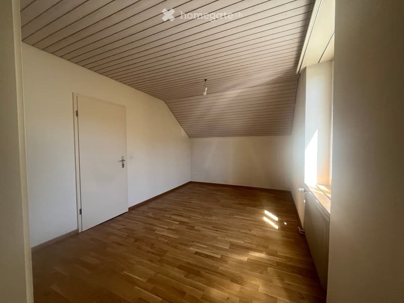 Empty room with wooden flooring, white walls, and a slanted ceiling. Features a single door and a window letting in natural light.