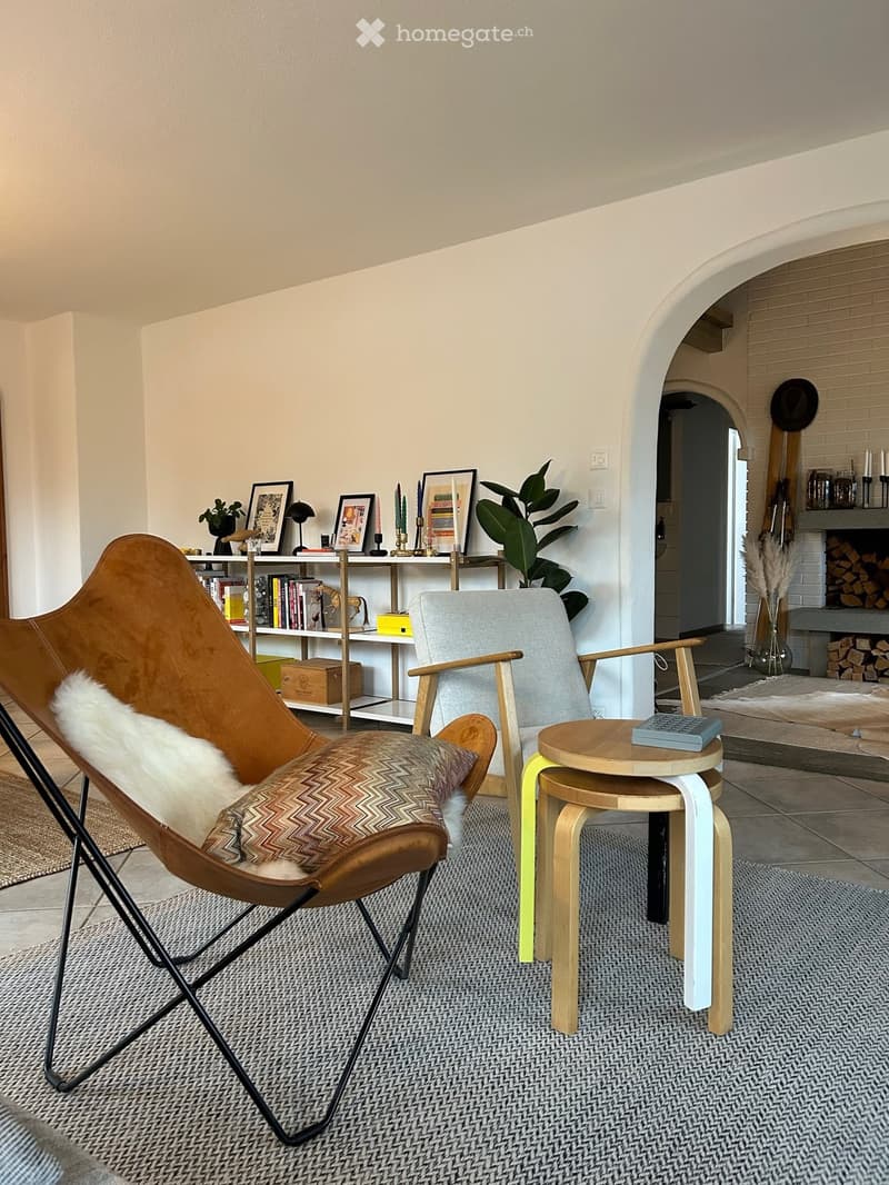 A modern living room with a brown leather chair, a grey armchair, a small wooden table, a shelf with books and decorative items, and a fireplace in the background.