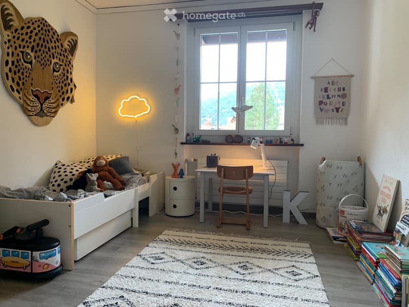 A cozy children's bedroom with a small sofa, desk, chair, and various toys and books. Features a large window with a view, a decorative leopard head wall hanging, and a colorful alphabet banner.