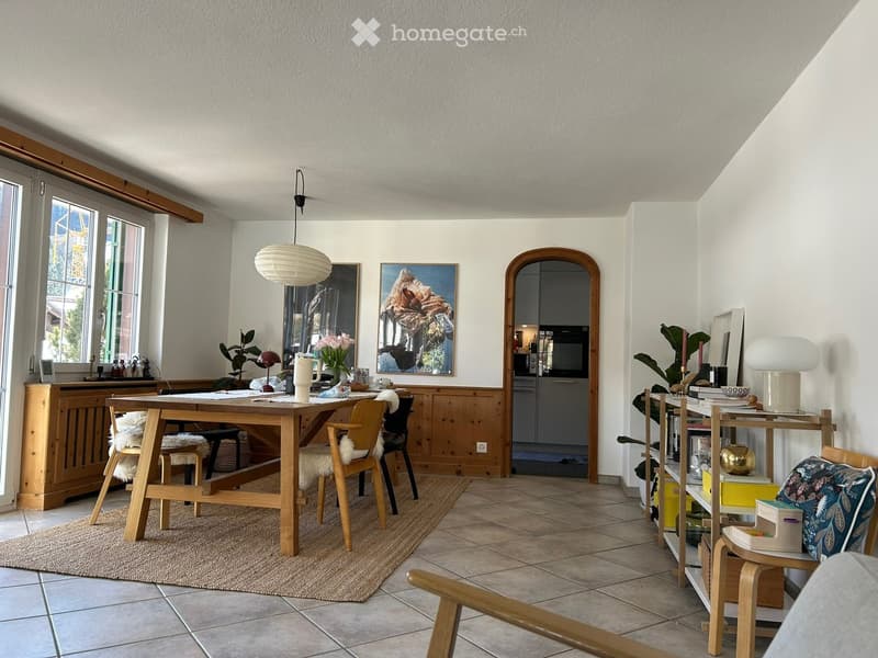 Bright, open room with a wooden dining table, chairs, shelves, and artwork. Features large windows, a hanging lamp, and a cozy rug.