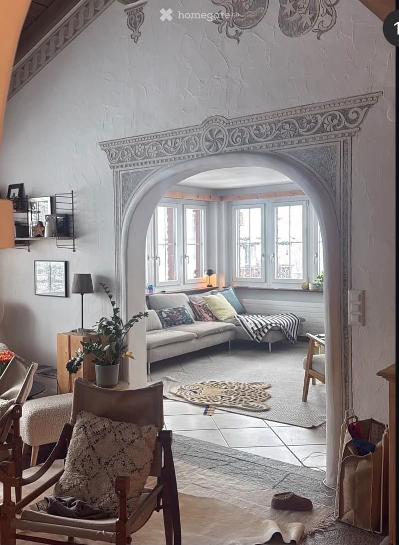 A cozy living room with a grey sofa, wooden chair, small table, lamp, and a potted plant. The room features a decorative arched doorway and a patterned rug on the floor.