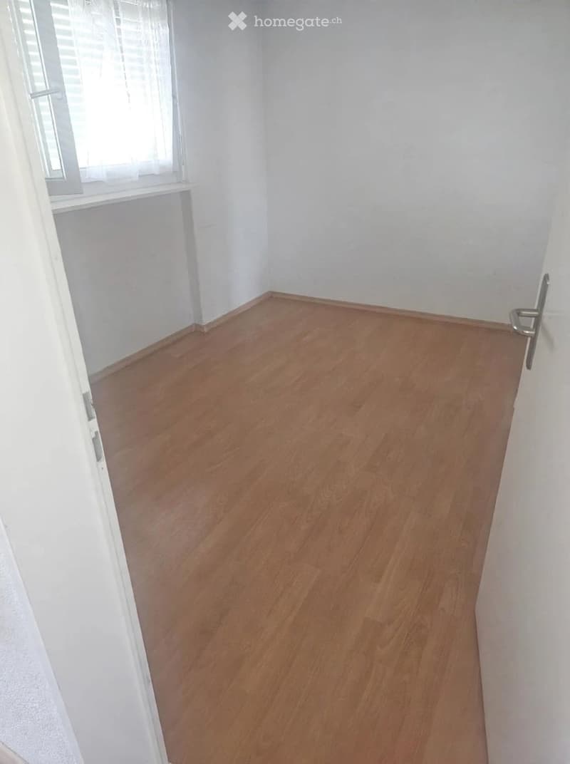 Empty room with wooden flooring and white walls, featuring a window with blinds and a door.