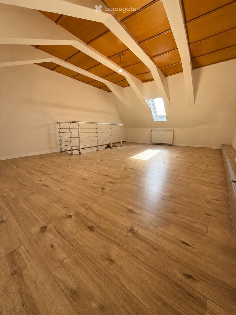 Empty attic space with wooden flooring, exposed wooden beams, and a metal railing leading to a small window. The room is well-lit with natural light coming through the window.