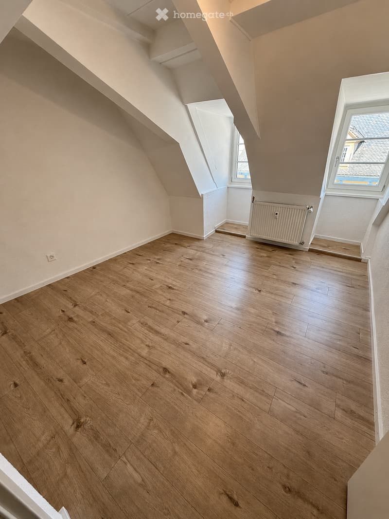 Empty attic room with wooden flooring, white walls, and two windows allowing natural light. The room features a slanted ceiling and a radiator on one wall.