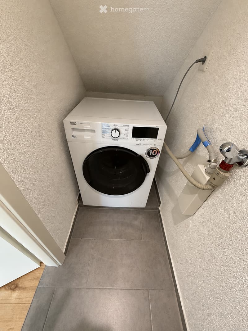A small, well-lit room with a white washing machine, tiled floor, and beige walls.