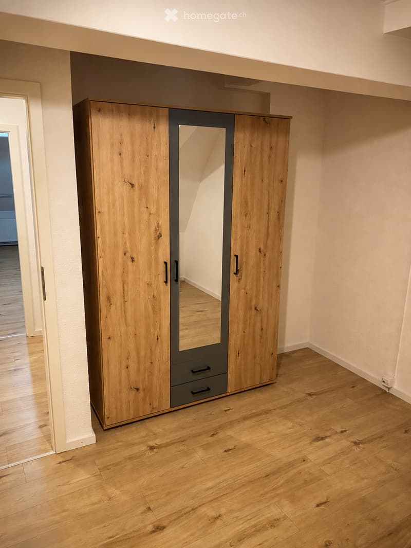 Empty room with wooden flooring, light-colored walls, and a large wooden wardrobe with mirrored doors and drawers.