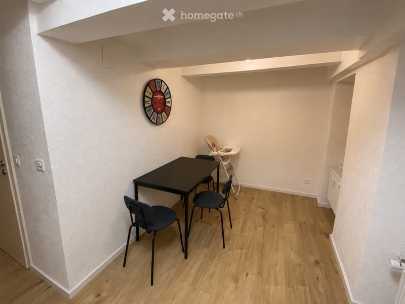 A small, furnished room with a black table, four chairs, a baby chair, and a wall-mounted clock. The room has light wooden flooring and white walls.
