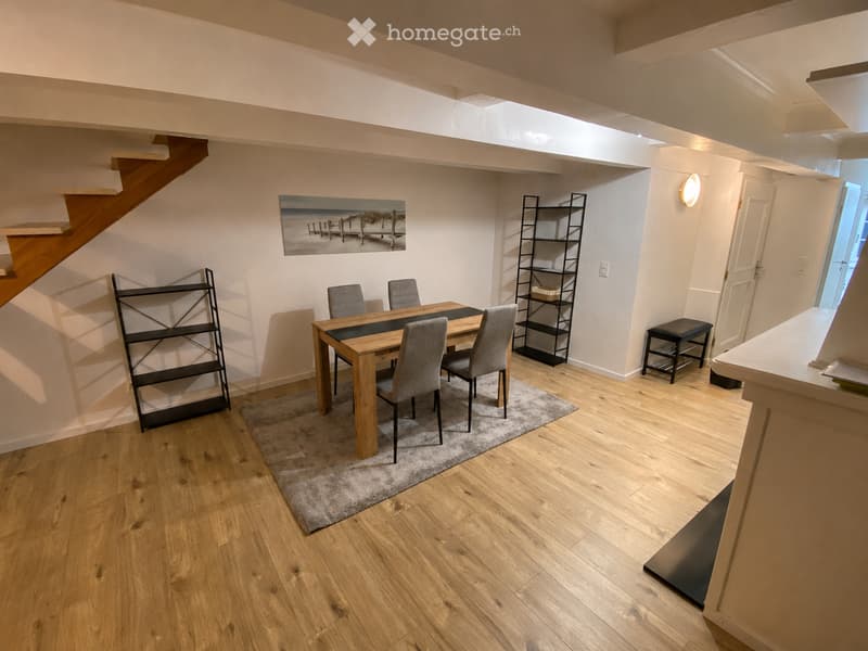 Dining area with wooden dining table, four chairs, two black shelves, a small stool, and a painting on the wall. Wooden flooring and a gray rug under the table.