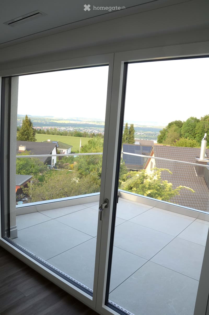Modern balcony with large glass doors, tiled floor, and expansive scenic view of countryside and houses.