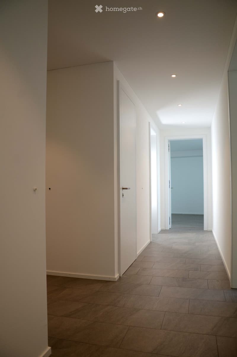 Empty hallway with tiled floor, white walls, and recessed ceiling lights. Features a closed door on the left and an open door leading to another room at the end.