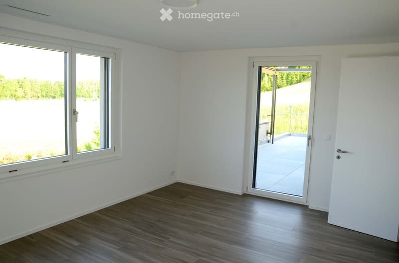 Empty room with white walls, wooden flooring, large windows, and a sliding glass door leading to a balcony.